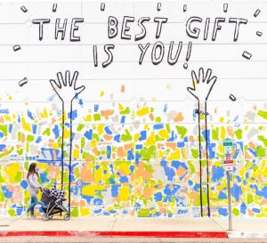 a lady pushing a pram past a wall with a multi-coloured mural. The mural features two raised hands and the words “The best gift is you!”