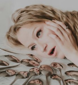 a woman is lying on her side looking tired.  she is facing an array of shiny metal spoons.  Her reflection is visible in each spoon.